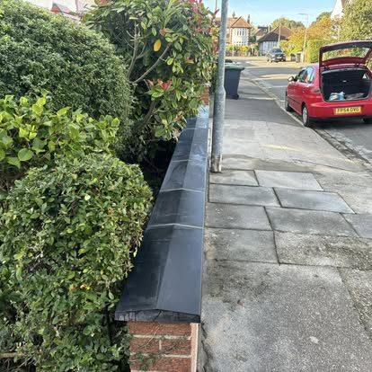 A low boundary wall with dark coping stones running along a pavement, with a hedge behind it and a parked car on the road