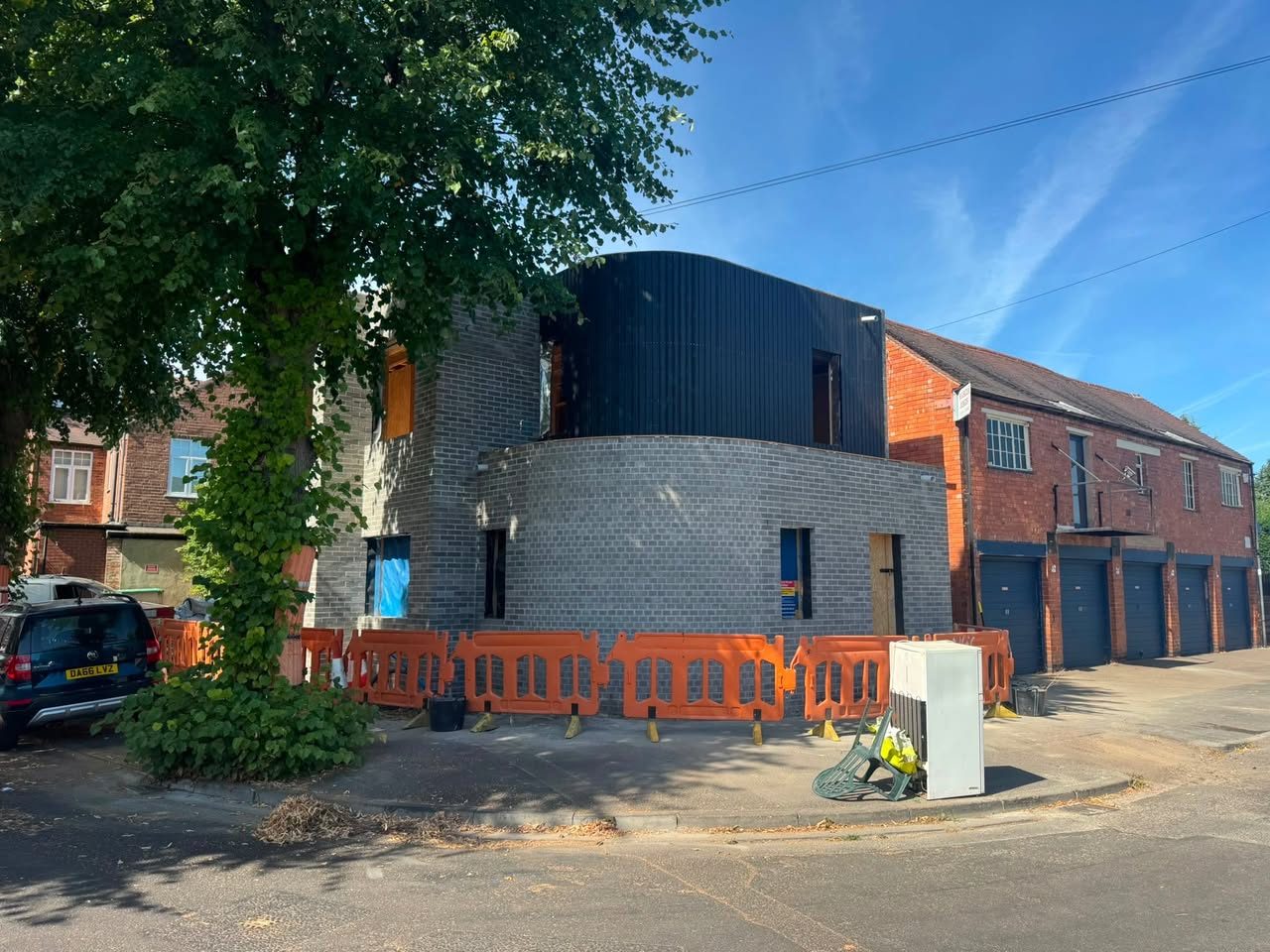 A curved grey brick wall on a new building, seen from across the street, with dark cladding above and orange barriers around the base
