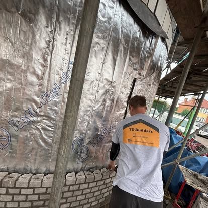 A bricklayer in a TD Builders t-shirt, photographed from behind, building the inside face of a curved wall against insulation sheeting
