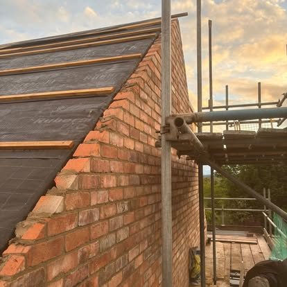 Red brickwork built up to a gable under scaffolding at sunset, with the timber roof structure resting on it